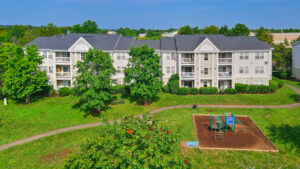 Aerial Exterior of Acclaim at Ashburn, neighborhoods in the background, lush foliage, neutral toned buildings in background, playground, photo taken on a sunny day.